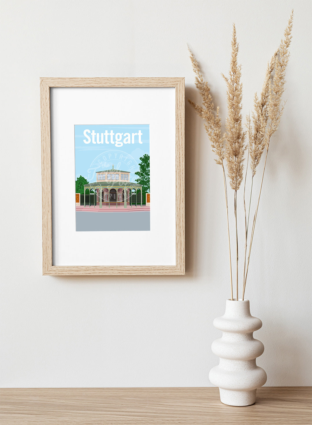 A frame with a postcard of the Stuttgart Wilhelma Kassenpavillon, hung on a white wall, above a wooden shelf, on which is a white ceramic vase containing reeds.