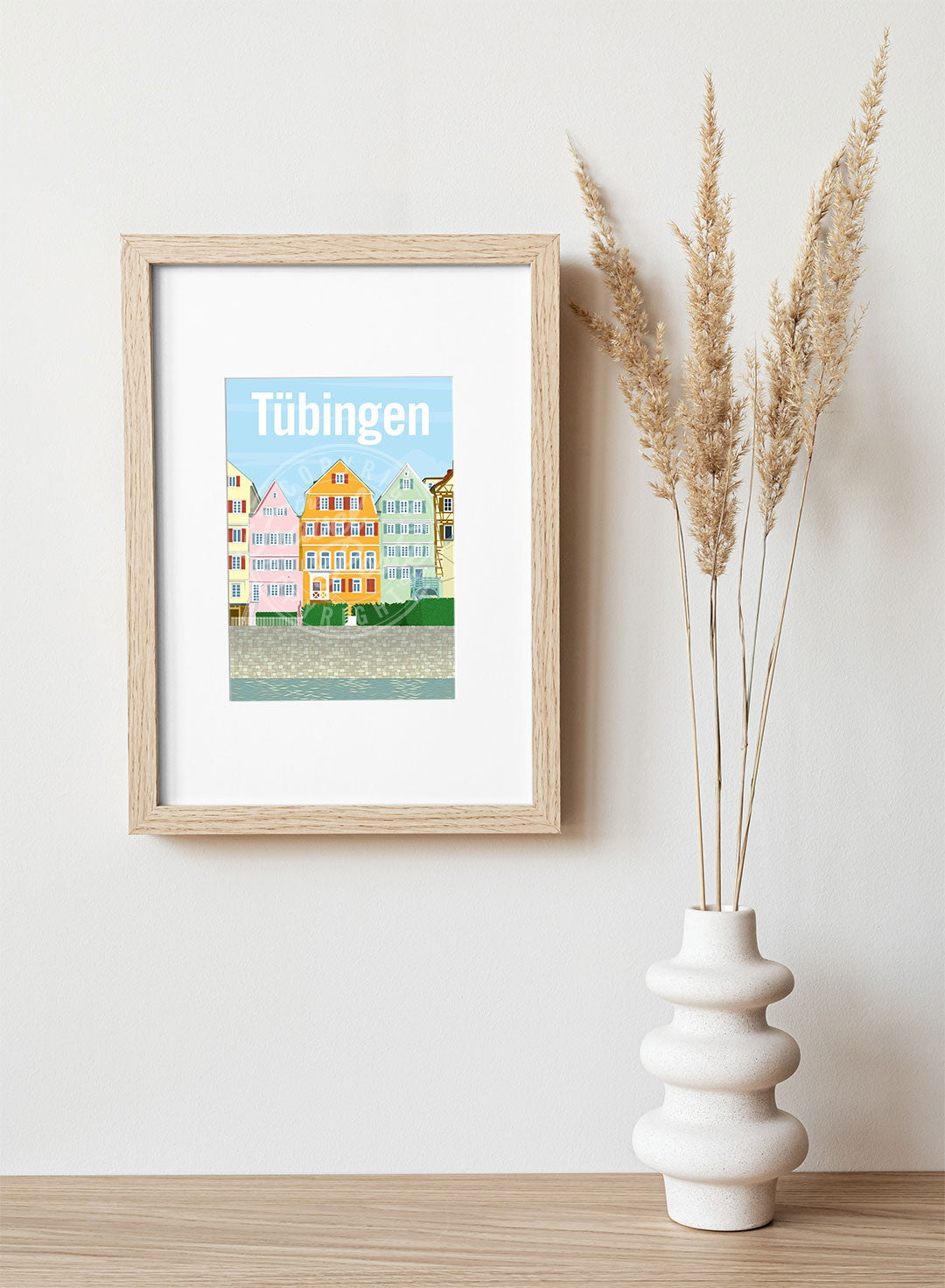 A frame with a postcard of the Tübingen Neckar front, hung on a white wall, above a wooden shelf, on which is a white ceramic vase containing reeds.