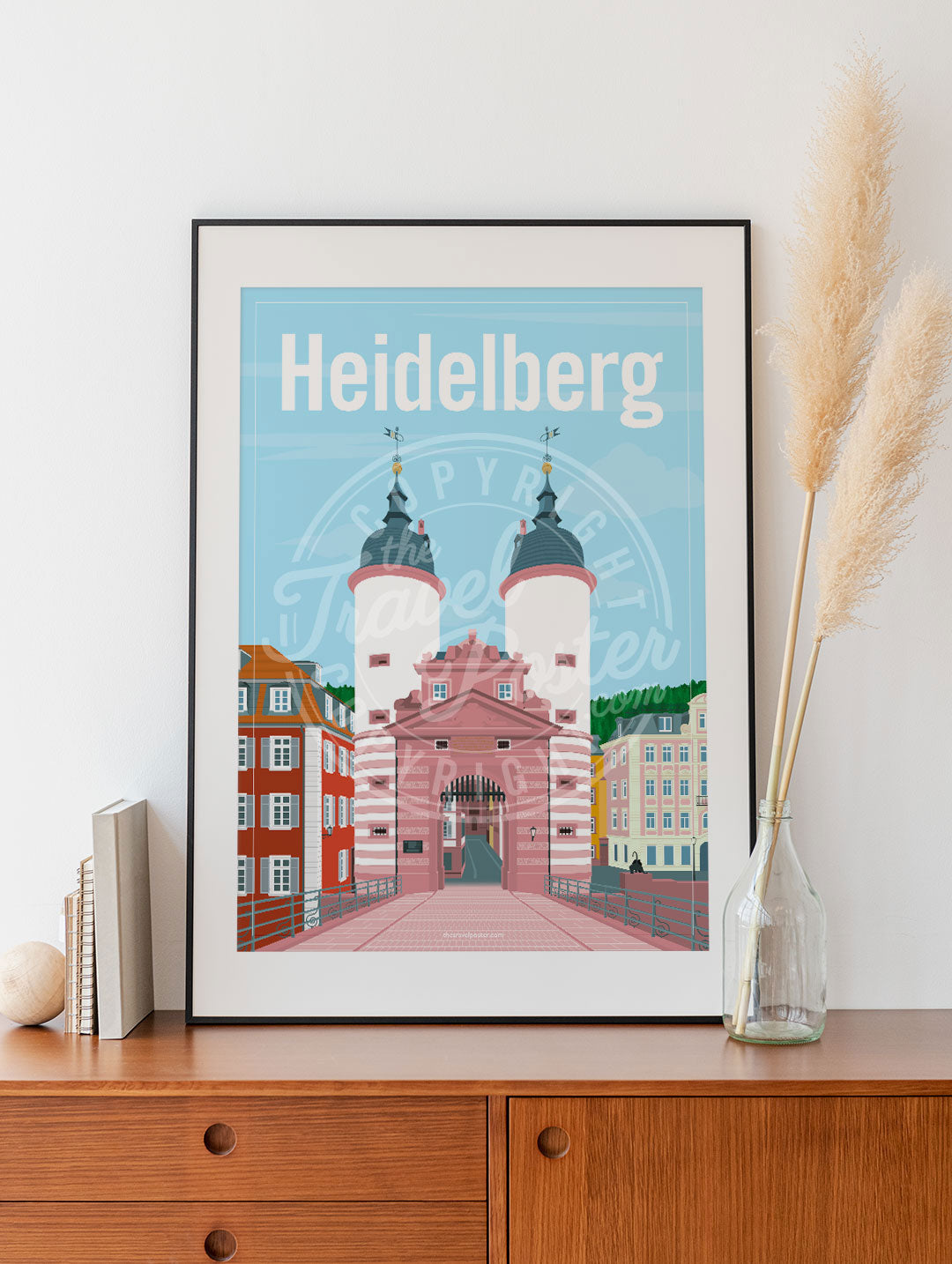 a frame with a poster of the Heidelberg Bridge Gate, placed on a wooden sideboard with some books and reeds in a glass bottle