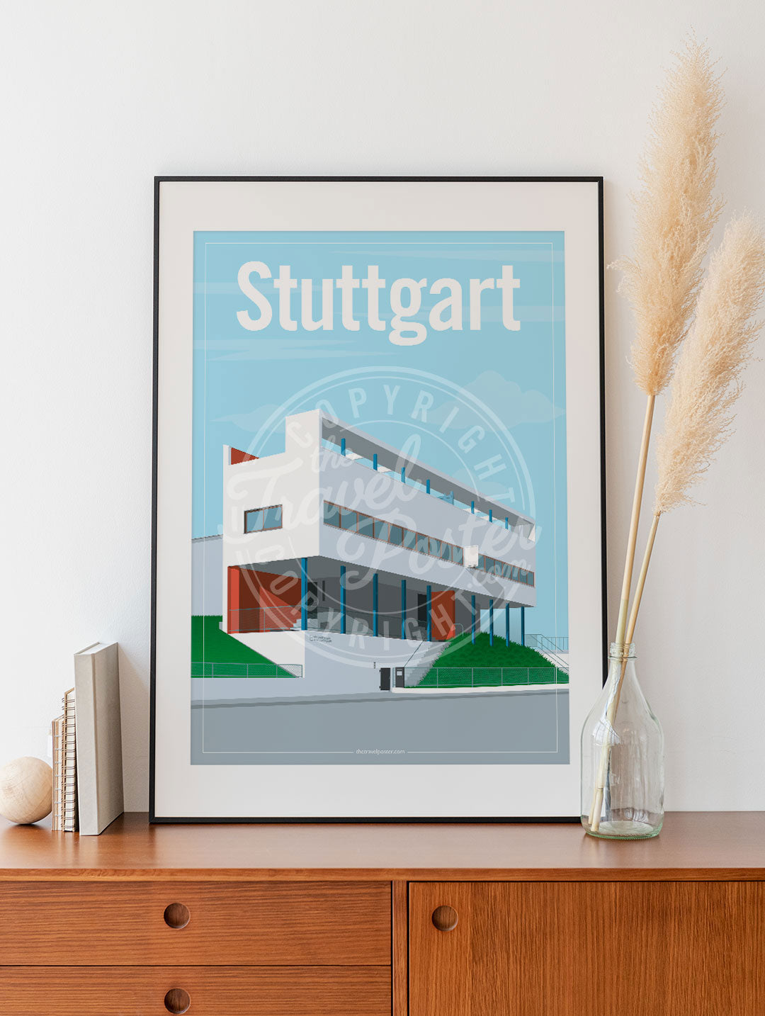a frame with a poster of the Stuttgart Weissenhof Museum, placed on a wooden sideboard with some books and reeds in a glass bottle