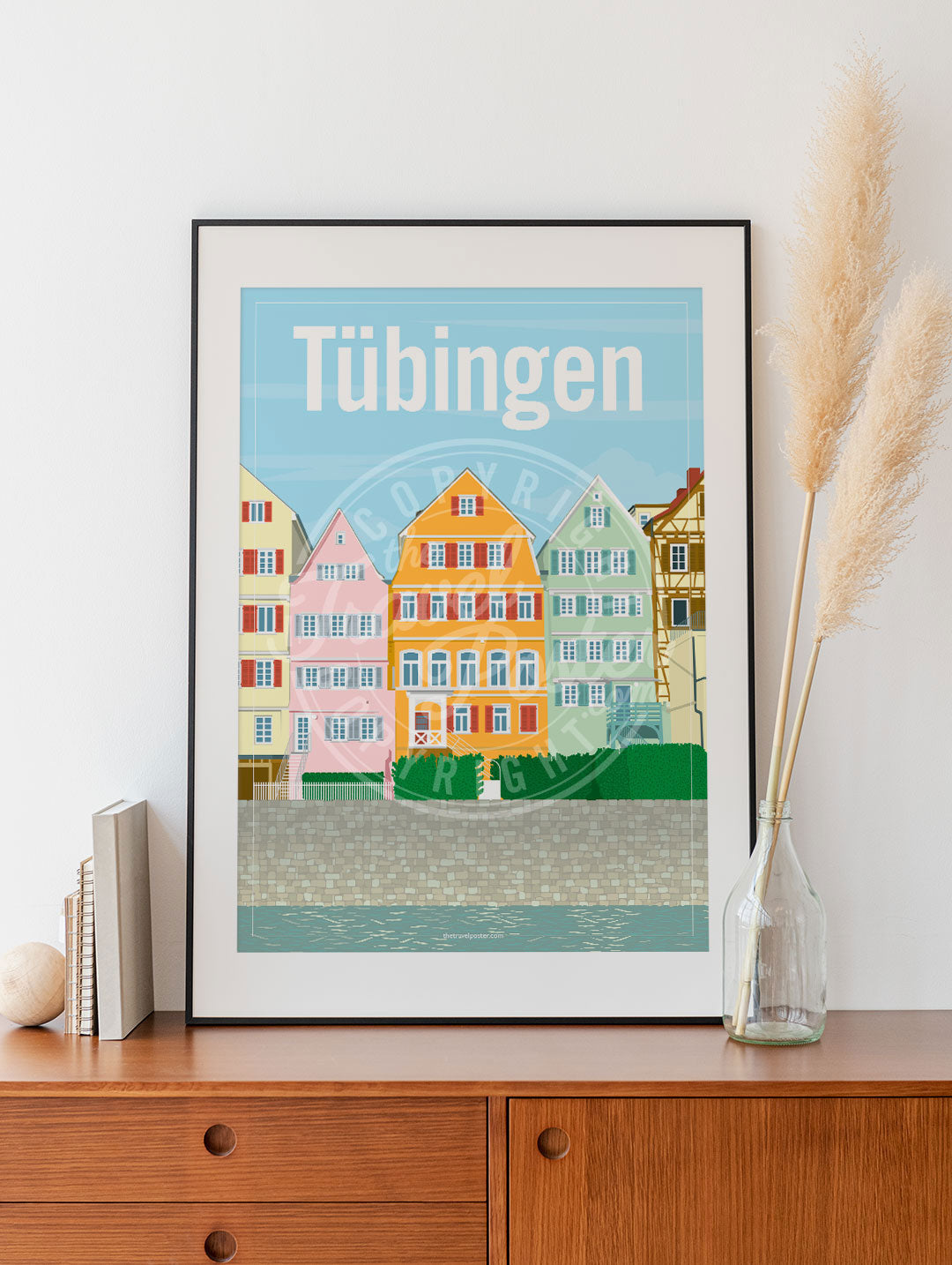 a frame with a poster of the colourful timber-framed houses on the Neckar riverfront in Tübingen, placed on a wooden sideboard with some books and reeds in a glass bottle