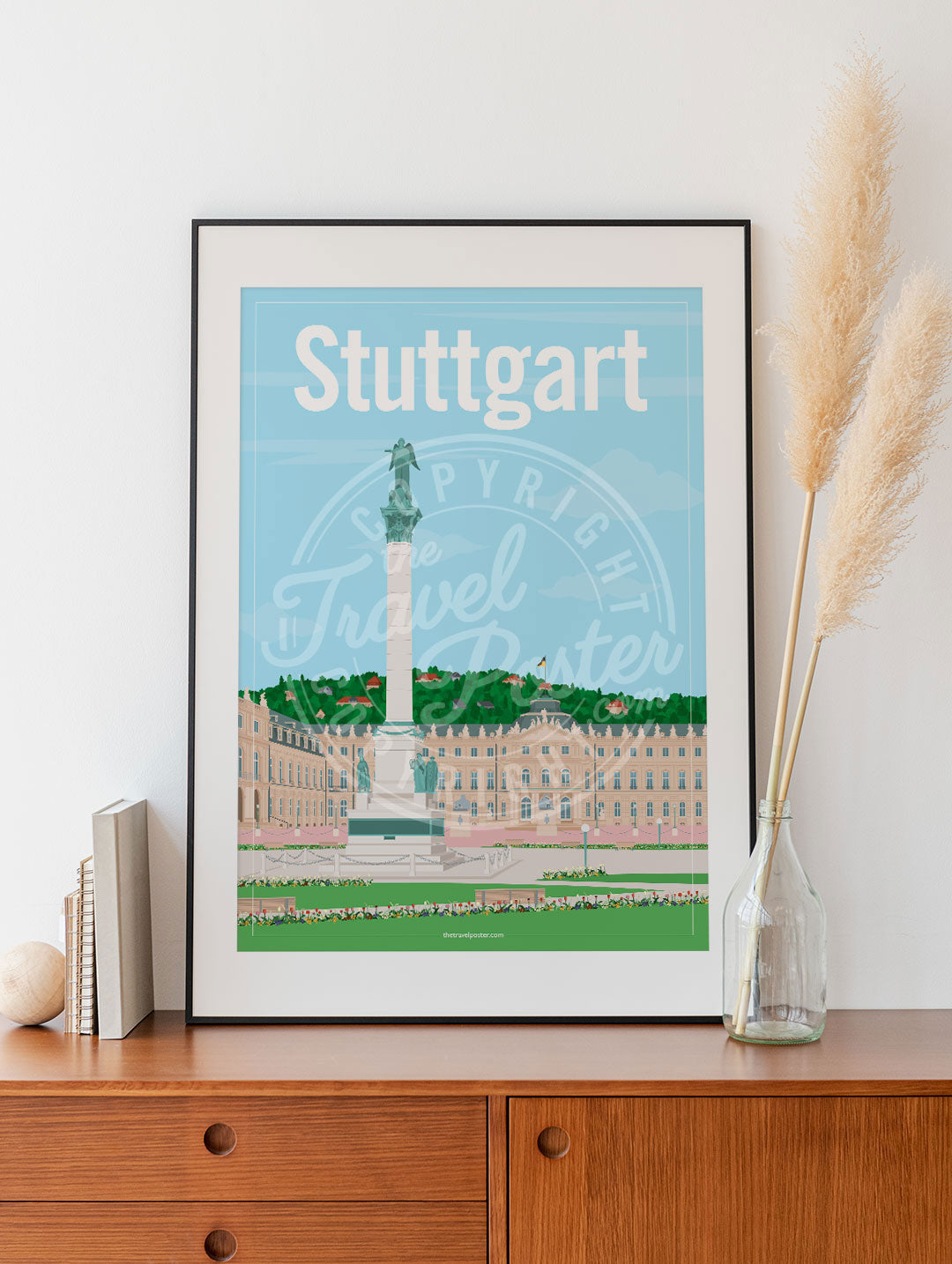 a frame with a poster of the Stuttgart Schlossplatz, placed on a wooden sideboard with some books and reeds in a glass bottle