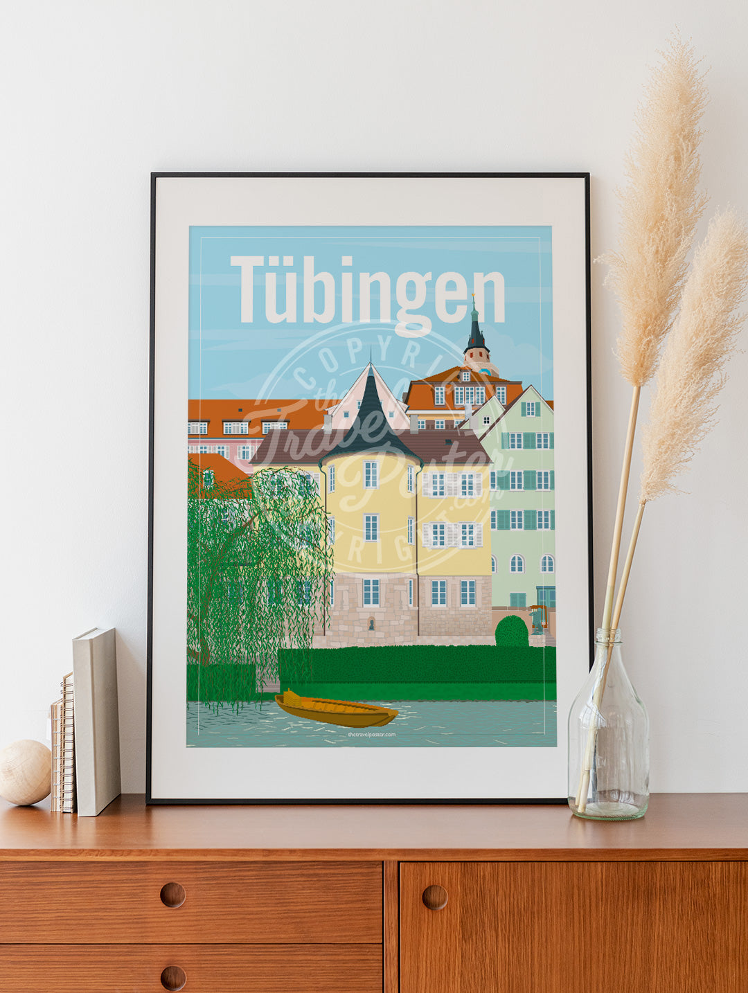 a frame with a poster of the Tübingen Holderlinturm, placed on a wooden sideboard with some books and reeds in a glass bottle