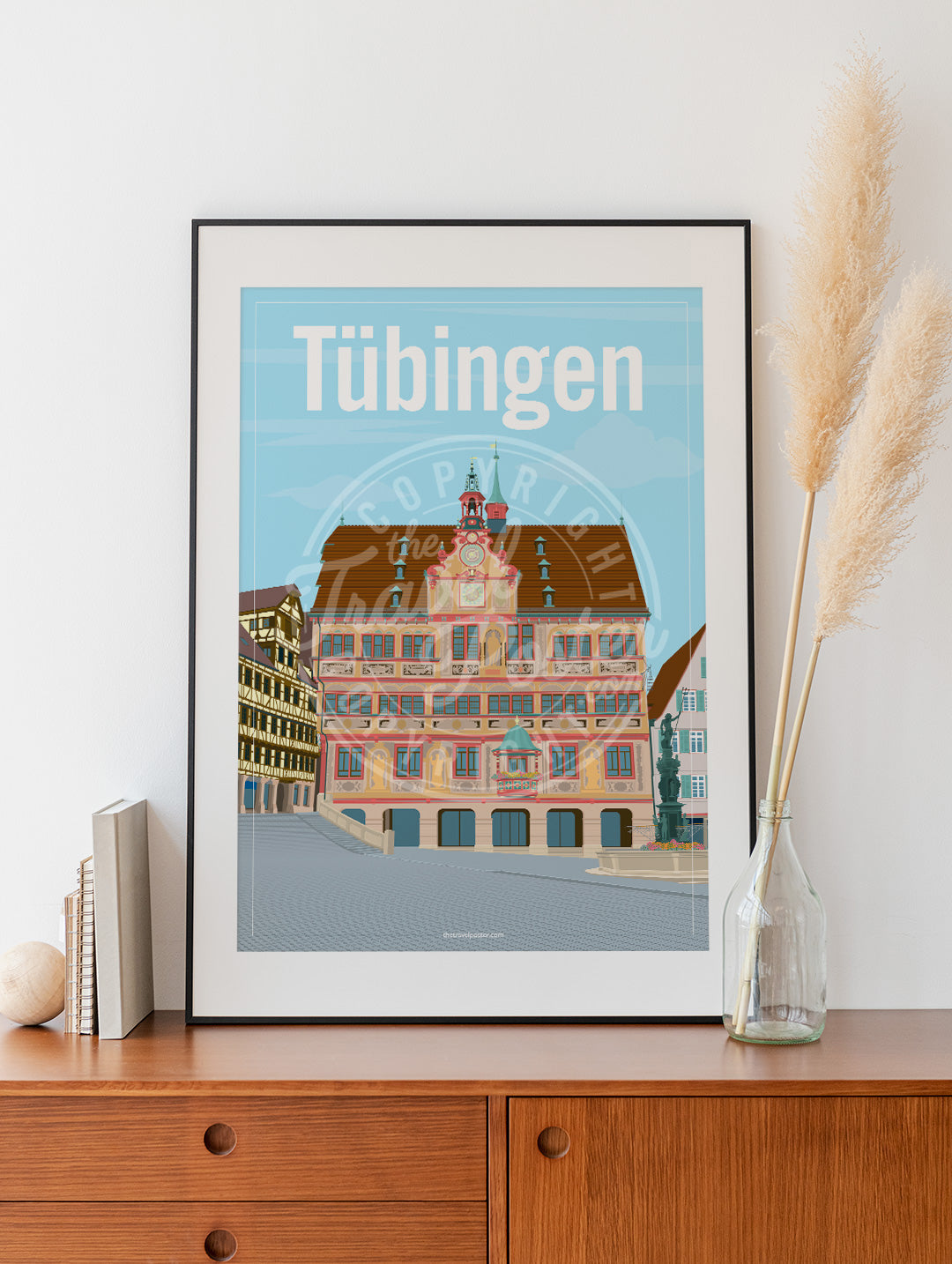 a frame with a poster of the Tübingen Rathaus, placed on a wooden sideboard with some books and reeds in a glass bottle