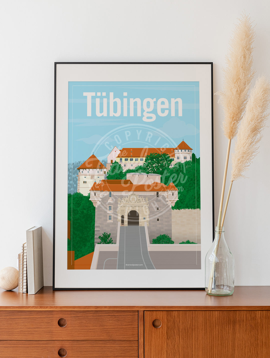a frame with a poster of the Hohentübingen Castle, placed on a wooden sideboard with some books and reeds in a glass bottle